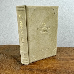 May include: A light tan hardcover book titled "The Princess Bride". The title and author's name, William Goldman, are embossed in gold. The book has a textured, suede-like cover. The book is in a vertical position.