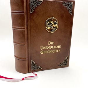 Può includere: Un libro rilegato in pelle marrone con il titolo tedesco "DIE UNENDLICHE GESCHICHTE" in rilievo dorato. Il libro presenta angoli decorativi in metallo e un emblema centrale. Un segnalibro a nastro rosso e bianco sporge dal libro.