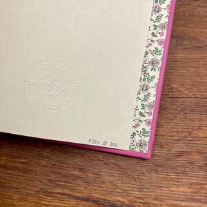 Puede incluir: Primer plano de un diario con una cubierta rosa y un borde con estampado floral. El diario tiene un logotipo en relieve y el texto "Unikat". La esquina inferior derecha tiene el texto "N&deg; 2559 AK 2026".