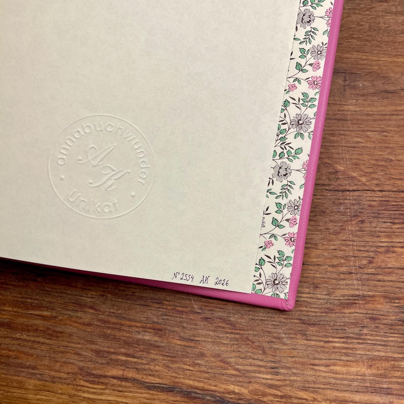 Puede incluir: Primer plano de un diario con una cubierta rosa y un borde con estampado floral. El diario tiene un logotipo en relieve y el texto "Unikat". La esquina inferior derecha tiene el texto "N&deg; 2559 AK 2026".