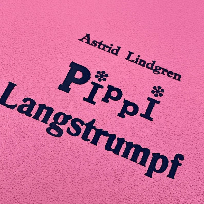 Puede incluir: Primer plano de una superficie rosa con el texto "Astrid Lindgren" y "Pippi Langstrumpf" en azul oscuro. El t&iacute;tulo est&aacute; en negrita, con acentos florales sobre las "P".