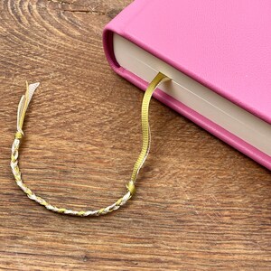 Puede incluir: Un diario rosa encuadernado en cuero con un marcap&aacute;ginas de cinta dorada. El marcap&aacute;ginas est&aacute; trenzado con hilos dorados y plateados. El diario est&aacute; sobre una superficie de madera.