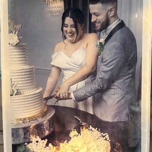 May include: A bride and groom smiling as they cut a white wedding cake with a silver knife. The cake is decorated with white frosting and pearl accents. The couple is standing in front of a table with a white floral arrangement.