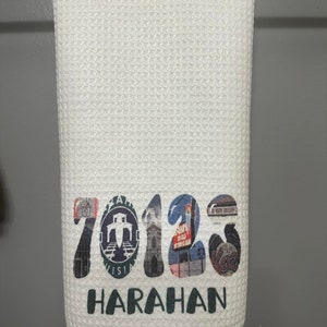 May include: White kitchen towel with a printed design of the numbers 2012 with images of buildings and a sign that says "Harahan".