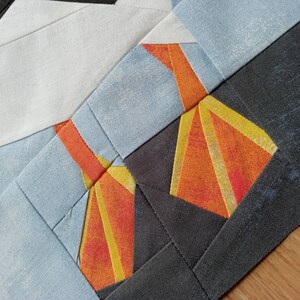 Puede incluir: Primer plano de un bloque de colcha con un dise&ntilde;o geom&eacute;trico. El bloque presenta una combinaci&oacute;n de telas naranja, amarilla y gris. La tela es texturizada y tiene un aspecto ligeramente desgastado.