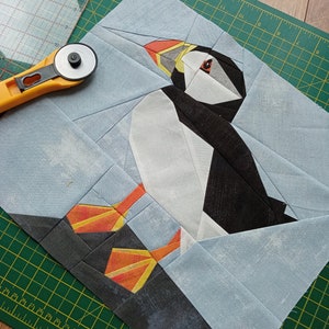 Puede incluir: Un mosaico de tela azul y blanca de un frailecillo con un pico naranja y ojos amarillos. El frailecillo est&aacute; de pie sobre un fondo de tela blanca y azul.