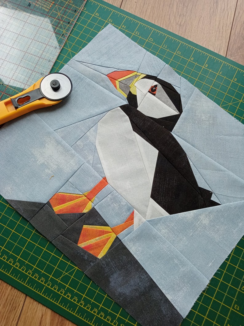 Puede incluir: Un mosaico de tela azul y blanca de un frailecillo con un pico naranja y ojos amarillos. El frailecillo est&aacute; de pie sobre un fondo de tela blanca y azul.
