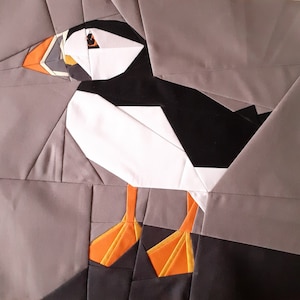 Puede incluir: Un origami de frailecillo hecho de tela, con plumaje blanco y negro y patas naranjas. El frailecillo est&aacute; sentado sobre un fondo gris.