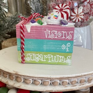 May include: A three-tiered wooden block set with the words "visions of sugarplums" painted on it. The top tier has a white frosting-like topping with colorful candies and a peppermint candy on top.