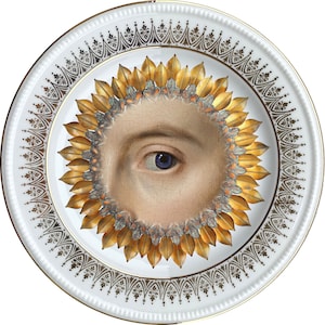 Op de afbeelding: Een wit porseleinen bord met een gouden en zwarte decoratieve rand. In het midden van het bord bevindt zich een enkel oog met een blauwe iris, omringd door een gouden zonnestralenontwerp.