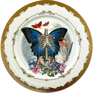 May include: A white ceramic plate with a gold rim features a blue and black butterfly with a skeletal body. The butterfly is surrounded by flowers and leaves in shades of red, pink, and green.