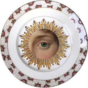 Op de afbeelding: Een wit porseleinen bord met een rode en witte bloemenrand. Het midden van het bord heeft een gouden en zilveren zonnestralenontwerp met een enkel oog in het midden.