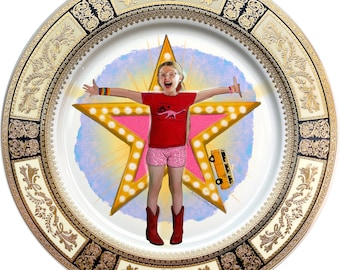 May include: A white ceramic plate with a gold and black decorative border. The center of the plate features a pink star with a girl standing in the middle. The girl is wearing a pink shirt with a dinosaur graphic and a pink skirt. The star has light bulbs around the edge. The text "AreliassoMadrid" is written in the bottom right corner of the plate.