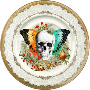 May include: A white porcelain plate with a gold rim and intricate white lace design. The center features a skull with a butterfly on each side, surrounded by red berries, pomegranates, and wheat.