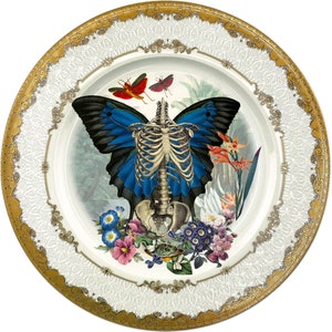 May include: A decorative plate with a gold rim and a white background features a blue butterfly with a skeletal body. The butterfly is surrounded by flowers and leaves in shades of red, pink, and green.