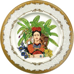 Puede incluir: Plato decorativo con un retrato de una mujer adornada con flores, rodeada de monos, loros y follaje exuberante. El plato tiene un centro blanco y un borde dorado y blanco.