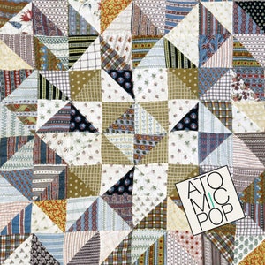 May include: A quilt made of various patterned fabrics in shades of brown, blue, white, and red. The quilt is made up of a repeating pattern of triangles, creating a star-like design in the center. The word "ATOMIC POP" is printed in white on a black rectangle in the lower right corner of the image.