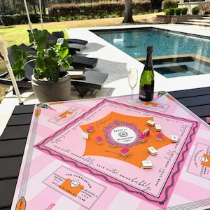 May include: A pink and orange Mahjong game board with the text "Press for Mahjong" and "Save water drink bubbly." A bottle of champagne and a glass of champagne are on the table. The game board is on a black table near a pool.