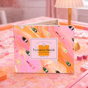 May include: A pink and orange marble-patterned box with champagne bottle illustrations. The box reads "Bird Bam Cheers" and "Veronica Smith Official Mahjong Card." Pink mahjong tiles, dice, and accessories are arranged on a pink surface.