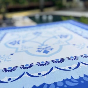 May include: Close-up of a blue and white patterned tablecloth. The design features floral and geometric motifs in shades of blue against a white background. The border is a darker shade of blue.