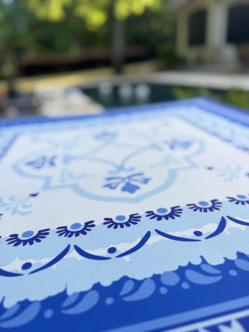 May include: Close-up of a blue and white patterned tablecloth. The design features floral and geometric motifs in shades of blue against a white background. The border is a darker shade of blue.