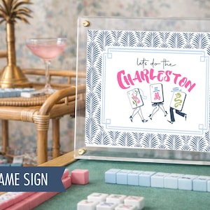 Puede incluir: Un letrero acr&iacute;lico transparente con herrajes dorados muestra la frase "Let's do the Charleston" en escritura rosa. El letrero tiene un borde con un patr&oacute;n de hojas azules y blancas. Fichas de Mahjong y un c&oacute;ctel en el fondo.