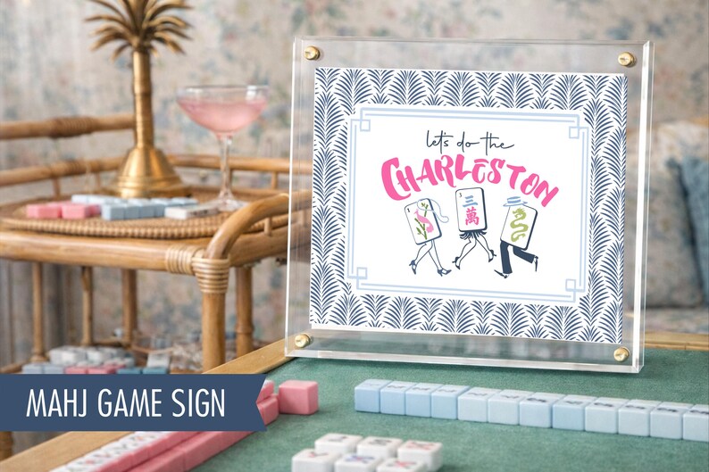 Puede incluir: Un letrero acr&iacute;lico transparente con herrajes dorados muestra la frase "Let's do the Charleston" en escritura rosa. El letrero tiene un borde con un patr&oacute;n de hojas azules y blancas. Fichas de Mahjong y un c&oacute;ctel en el fondo.
