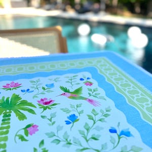 May include: A close-up of a light blue and white decorative table mat with a floral and hummingbird design. The mat features pink and blue flowers, green leaves, and a palm tree illustration. The border is light blue and green.
