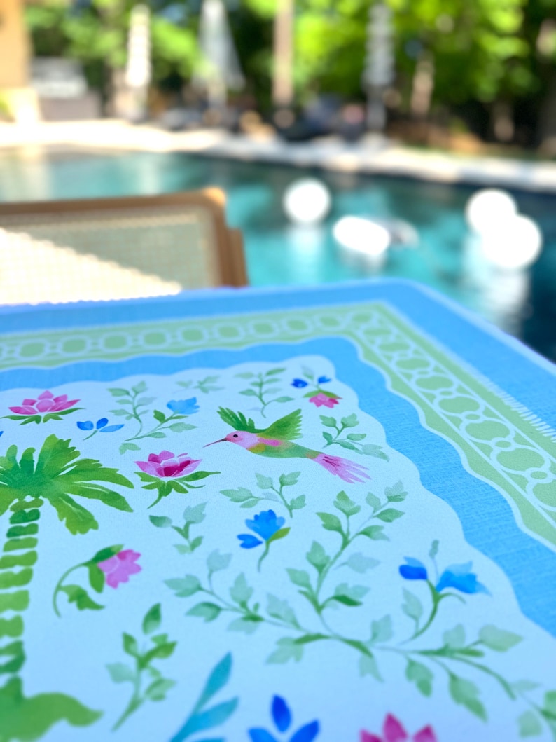 May include: A close-up of a light blue and white decorative table mat with a floral and hummingbird design. The mat features pink and blue flowers, green leaves, and a palm tree illustration. The border is light blue and green.