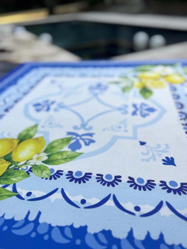 K&ouml;nnte beinhalten: Ein rechteckiger Tischset mit einem wei&szlig;en und blauen Blumendesign. Das Tischset zeigt ein Muster aus Zitronen, Bl&auml;ttern und Blumen. Der Rand ist tiefblau mit einem gewellten Rand.