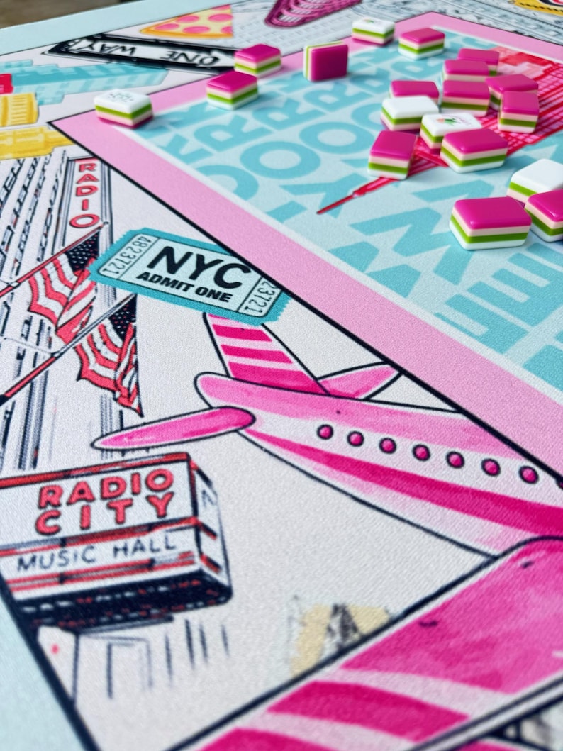 May include: A board game with a New York City theme. The game board features a pink airplane, the Radio City Music Hall, and an NYC admit one ticket. Pink, white, and green game pieces are scattered across a light blue and pink game board.