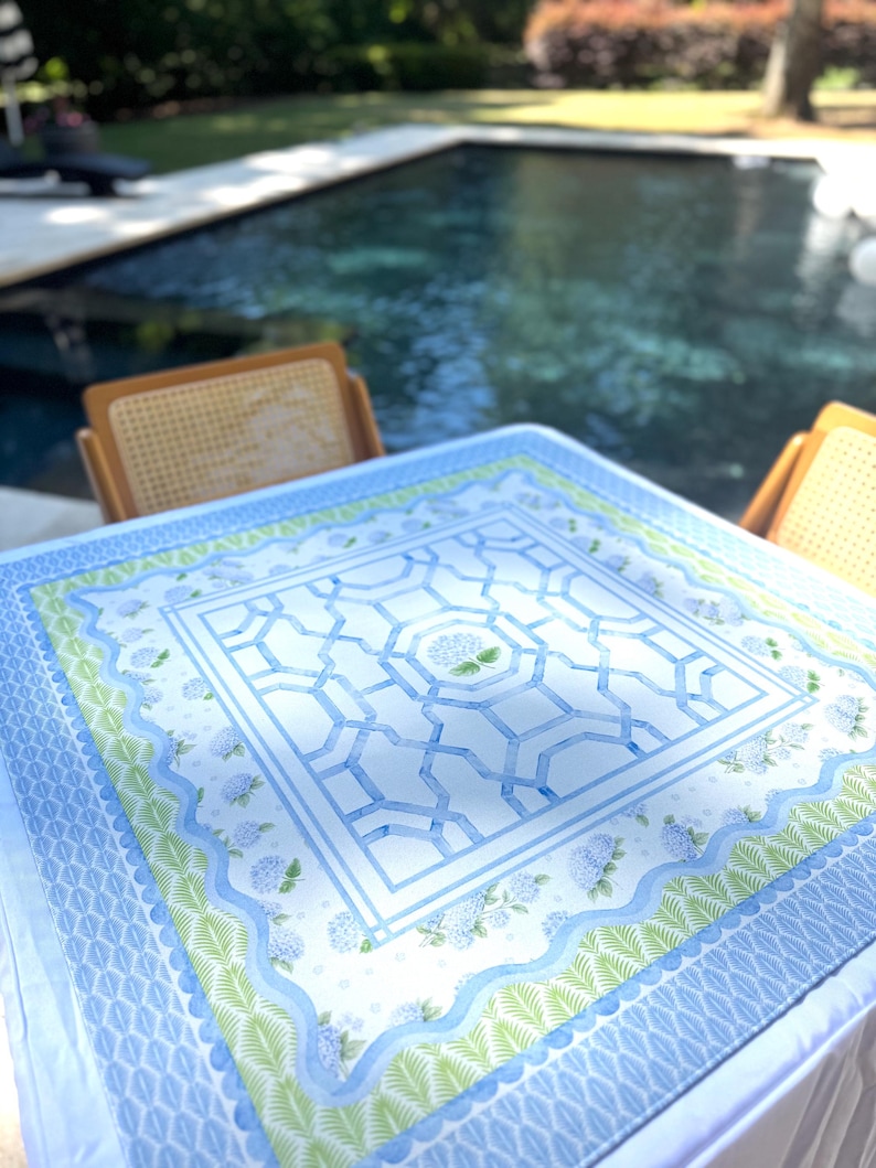 May include: A square tablecloth with a blue and white geometric design, featuring a central pattern and floral accents. The border includes a scalloped edge with green and blue floral and leaf patterns. The tablecloth is set on a table near a pool.