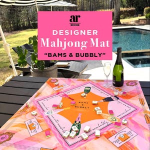 May include: A designer Mahjong mat with a pink and orange color scheme, featuring the text "BAMS & BUBBLY". The mat is set on a dark wooden table, with Mahjong tiles and a bottle of champagne. An outdoor setting with an umbrella and pool is visible.