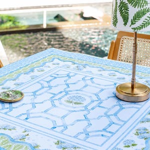 May include: A table setting featuring a light blue tablecloth with a geometric pattern and floral accents. A small brass lamp with a fern-patterned shade sits on the table, along with a small decorative dish. Two wooden chairs with woven backs are visible.