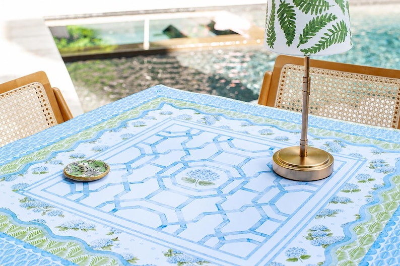 May include: A table setting featuring a light blue tablecloth with a geometric pattern and floral accents. A small brass lamp with a fern-patterned shade sits on the table, along with a small decorative dish. Two wooden chairs with woven backs are visible.