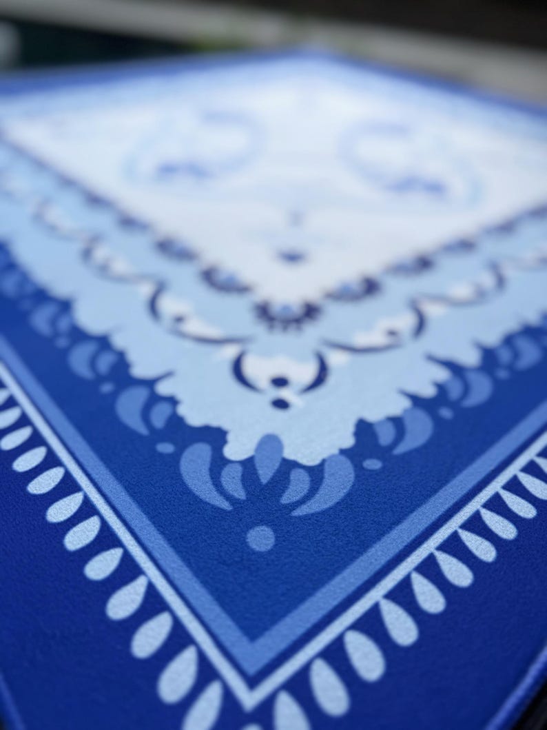 May include: Close-up of a blue and white patterned textile. The design features a central square with a floral motif, surrounded by borders of varying shades of blue. The corners have a teardrop-shaped pattern. The textile has a textured appearance.