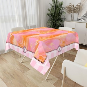 May include: A rectangular tablecloth with a pink and orange marble design. The tablecloth has a white and pink striped border with a decorative emblem. The tablecloth is set on a folding table.