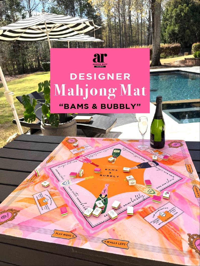 May include: A designer Mahjong mat with a pink and orange color scheme, featuring the text "BAMS & BUBBLY". The mat is set on a dark wooden table, with Mahjong tiles and a bottle of champagne. An outdoor setting with an umbrella and pool is visible.