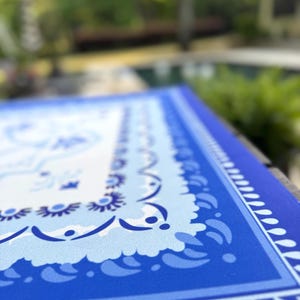 May include: A close-up of a blue and white patterned table runner. The runner features a detailed border design with a repeating pattern of teardrop shapes and floral motifs. The color palette includes shades of blue and white.
