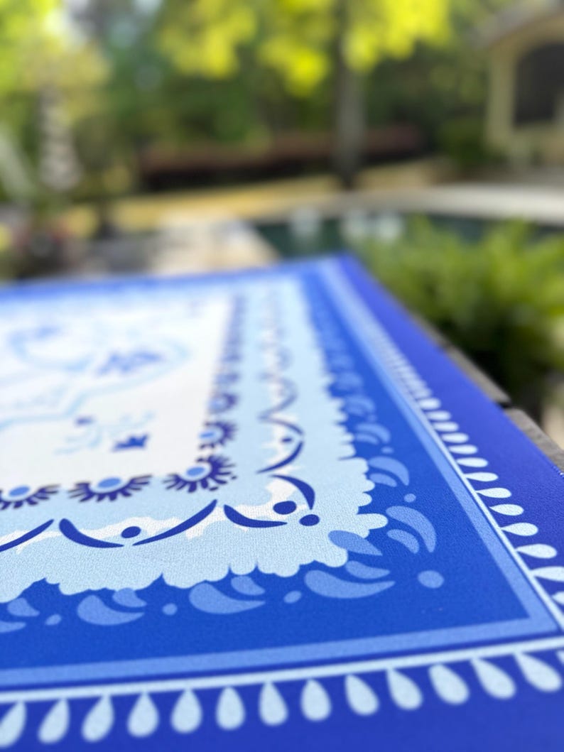 May include: A close-up of a blue and white patterned table runner. The runner features a detailed border design with a repeating pattern of teardrop shapes and floral motifs. The color palette includes shades of blue and white.