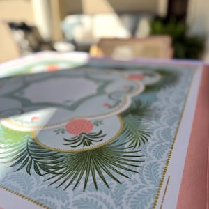 May include: Close-up of a decorative journal or notebook with a pink cover. The interior pages feature a light blue background with a repeating pattern and botanical illustrations in green and pink. Gold accents frame the design.