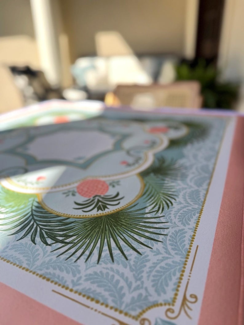 May include: Close-up of a decorative journal or notebook with a pink cover. The interior pages feature a light blue background with a repeating pattern and botanical illustrations in green and pink. Gold accents frame the design.