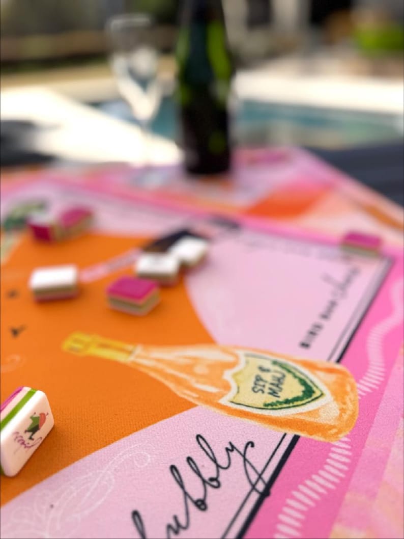 May include: A colorful table setting with a pink, orange, and white design. The table features a champagne bottle illustration with the words "SIP & MAU" and the word "bubbly". Small, rectangular objects with pink and white accents are scattered across the table.