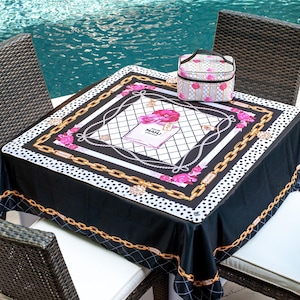 May include: A black tablecloth with a white and gold chain design, pink floral accents, and the text "MAHI" on a book. A matching patterned cosmetic bag sits on the table. The table is surrounded by four wicker chairs.