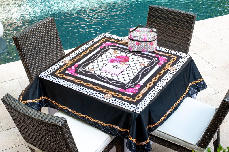 May include: A black tablecloth with a white and gold chain design, pink floral accents, and the text "MAHI" on a book. A matching patterned cosmetic bag sits on the table. The table is surrounded by four wicker chairs.