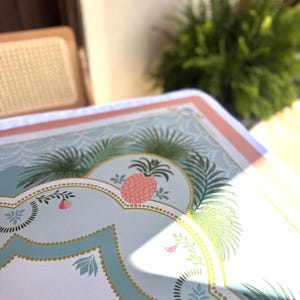 May include: A decorative serving tray with a light blue background and a pink and gold border. The tray features a pink pineapple design and green palm fronds. The tray is likely used for serving food or drinks.