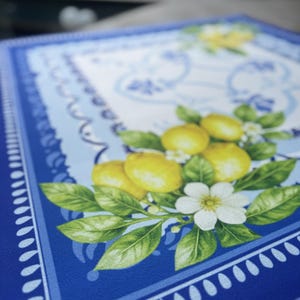 K&ouml;nnte beinhalten: Ein quadratischer, blauer Tischset mit dekorativem Rand und einem zentralen Design mit Zitronen, gr&uuml;nen Bl&auml;ttern und wei&szlig;en Bl&uuml;ten. Die Zitronen sind gelb und die Bl&auml;tter sind leuchtend gr&uuml;n.