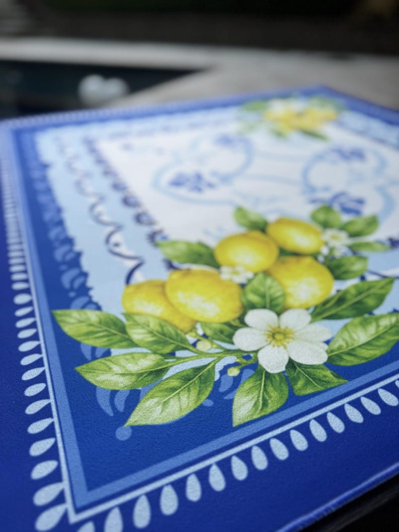 K&ouml;nnte beinhalten: Ein quadratischer, blauer Tischset mit dekorativem Rand und einem zentralen Design mit Zitronen, gr&uuml;nen Bl&auml;ttern und wei&szlig;en Bl&uuml;ten. Die Zitronen sind gelb und die Bl&auml;tter sind leuchtend gr&uuml;n.