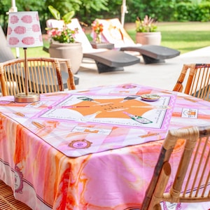 May include: A square table covered with a pink and orange patterned tablecloth, surrounded by four bamboo chairs. A small lamp with a red and white patterned shade sits on the table. The table has a board game on it.