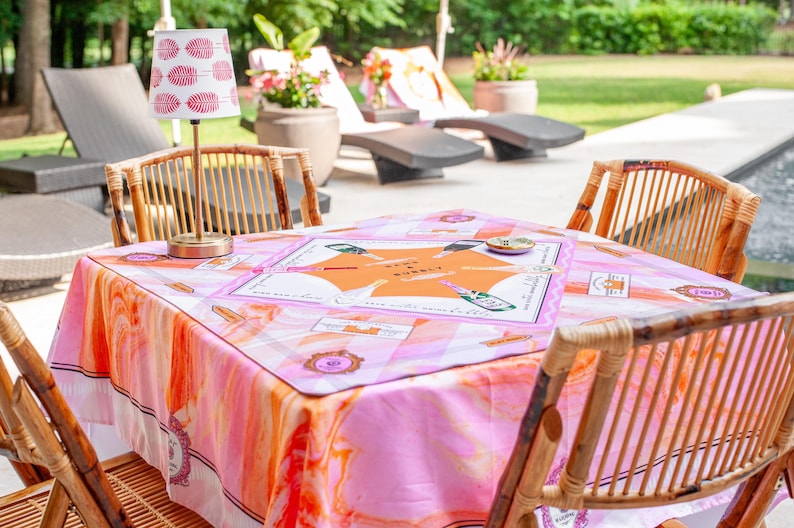 May include: A square table covered with a pink and orange patterned tablecloth, surrounded by four bamboo chairs. A small lamp with a red and white patterned shade sits on the table. The table has a board game on it.